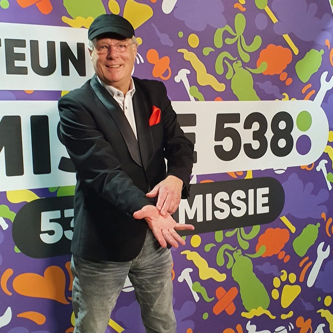 Professor Drabb in zwart colbert met rode pochet wijst naar zijn handpalm voor een kleurrijke fotowand van Missie 538