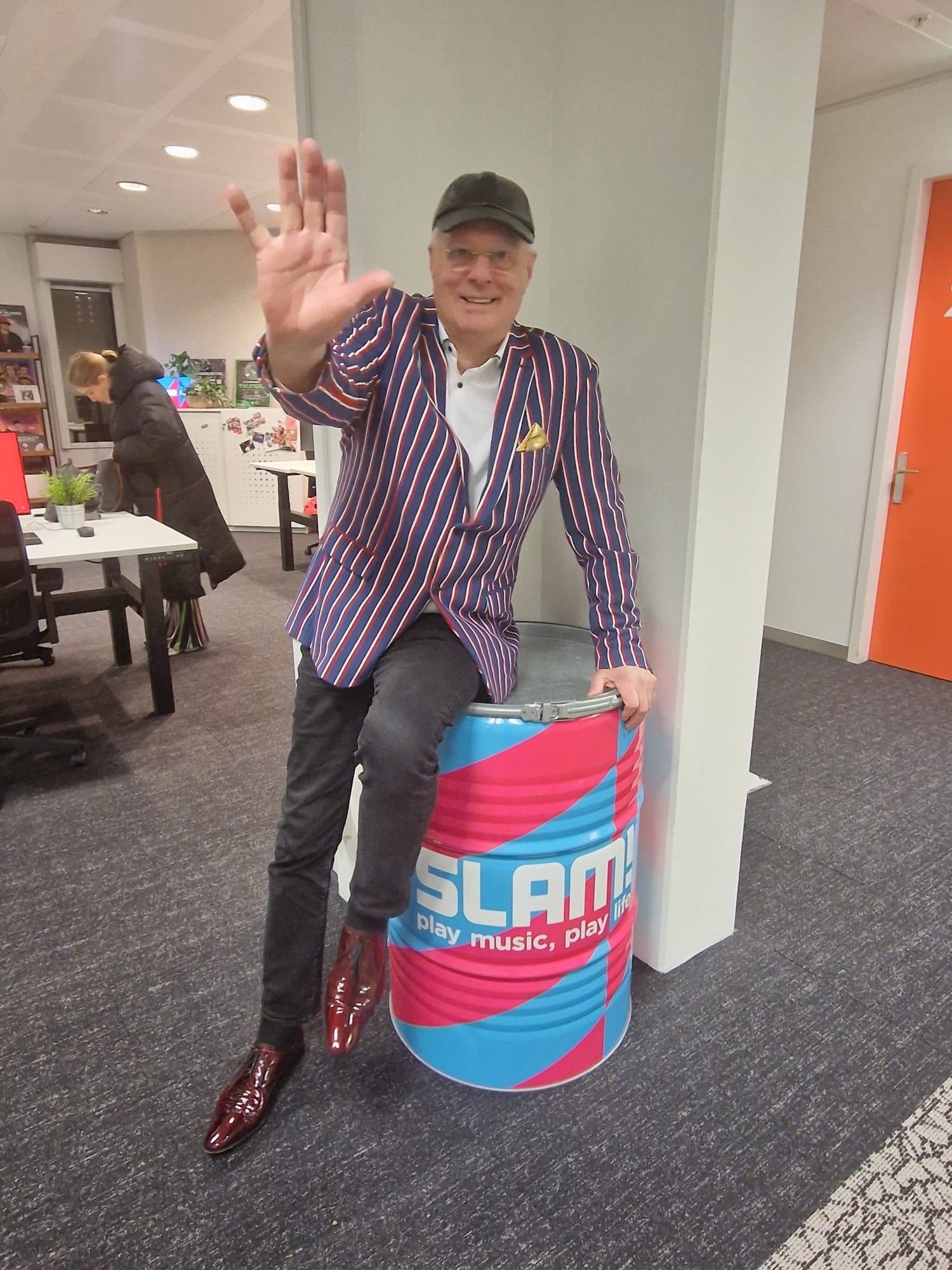 Professor Drabb in gestreept colbert zwaait vrolijk, zittend op een roze-blauwe SLAM!-ton op de redactie