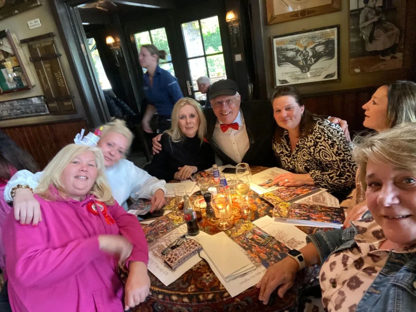 Professor Drabb met rode strik tussen een groep lachende vrouwen aan een ronde tafel in een café
